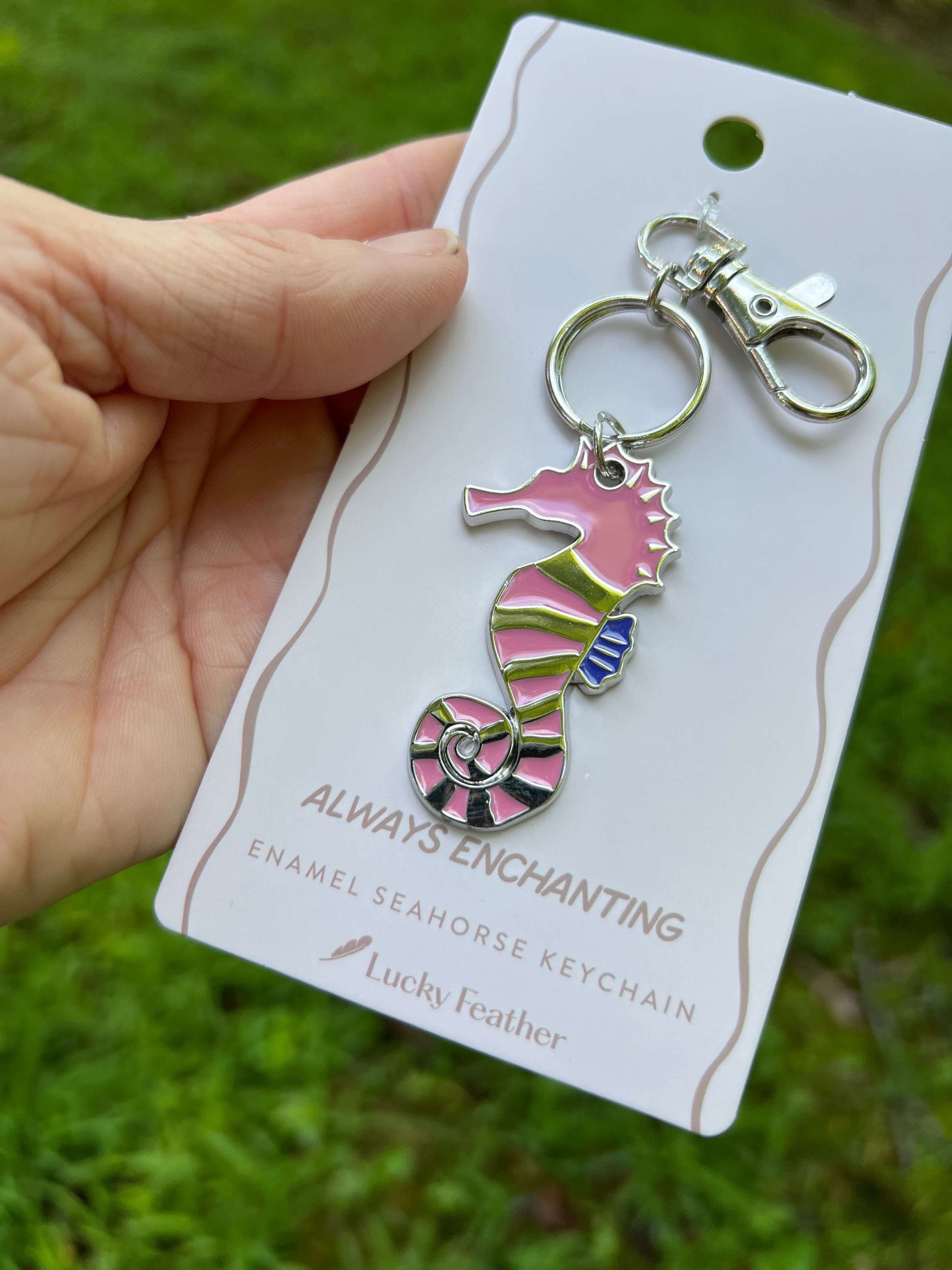 A hand holds a pink and green enamel seahorse keychain on a white branded card, with a blurred green grass background.