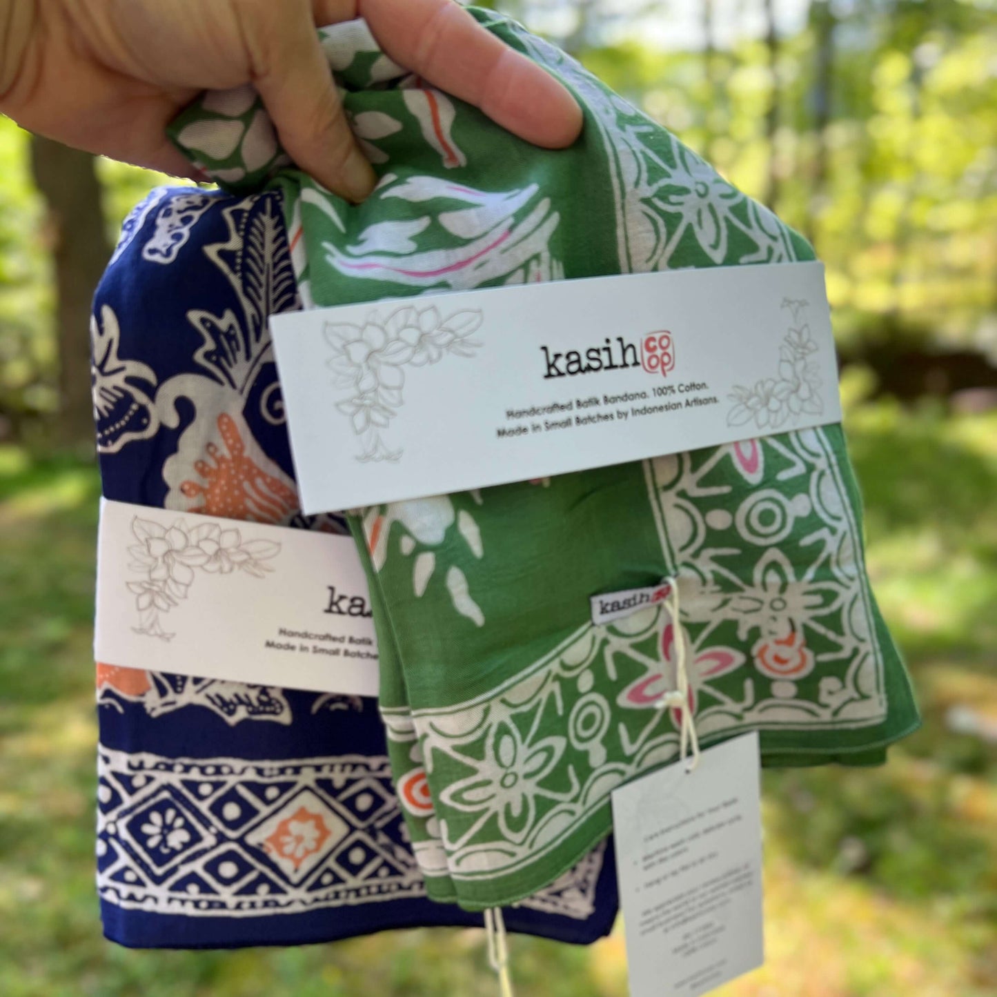 Hand holding a folded fabric product with 'kasih' branding against a blurred natural background