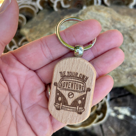 A hand holding a wooden dog tag keychain engraved with a retro VW bus and the words 'BE YOUR OWN ADVENTURE'. The keychain has a silver metal split ring and is photographed against a blurred natural background of rocks and moss.