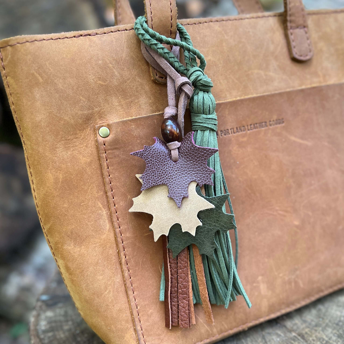 A close-up of a tan leather tote bag with a decorative hanging tassel keychain. The keychain features three layered leaf-shaped leather charms in purple, tan, and dark green, along with braided green cords and tassels.