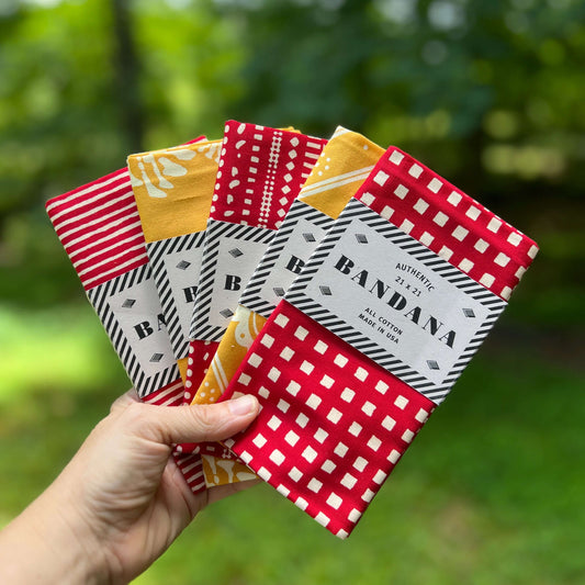 Hand holding a collection of patterned bandanas with 'B' brand label against a blurred green background