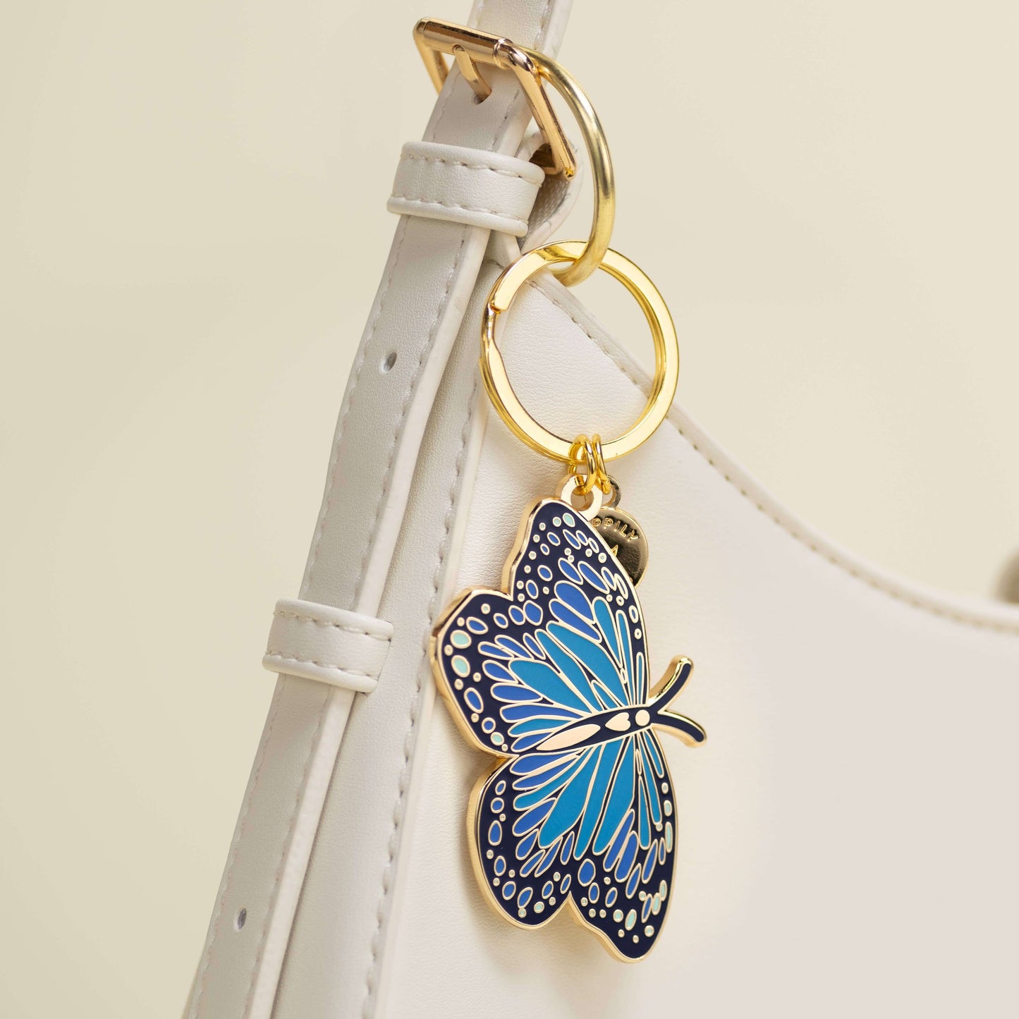 Blue butterfly keychain attached to a white strap with gold rings on a beige background
