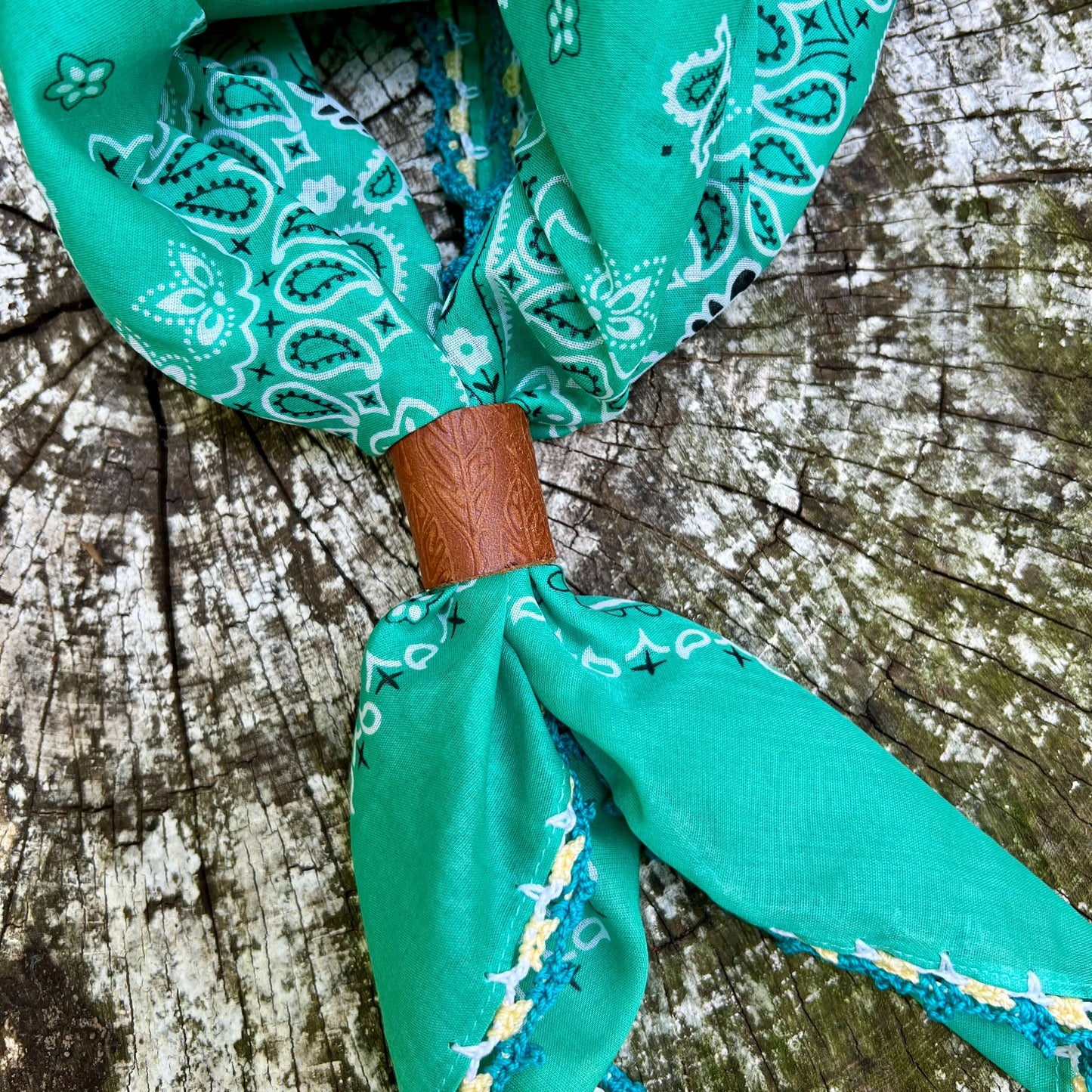 A green paisley bandana tie-on scarf with a brown leather wrap, displayed on a weathered wood surface.