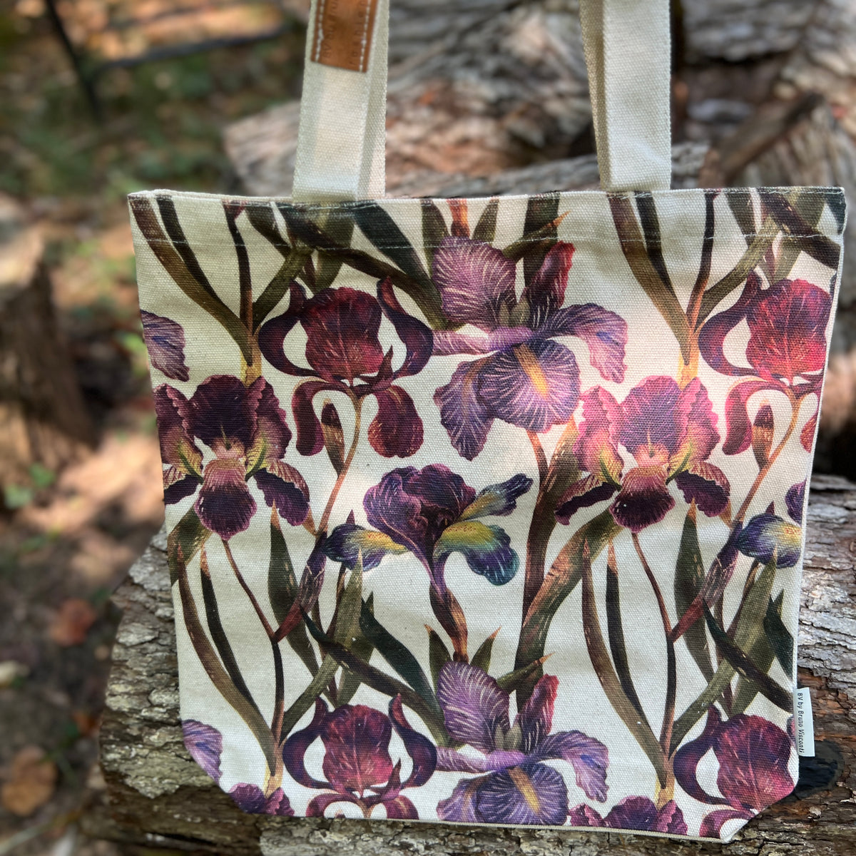 A canvas tote bag with a watercolor-style purple and burgundy iris floral print, featuring natural beige canvas handles, photographed outdoors on a tree trunk with a blurred forest background.