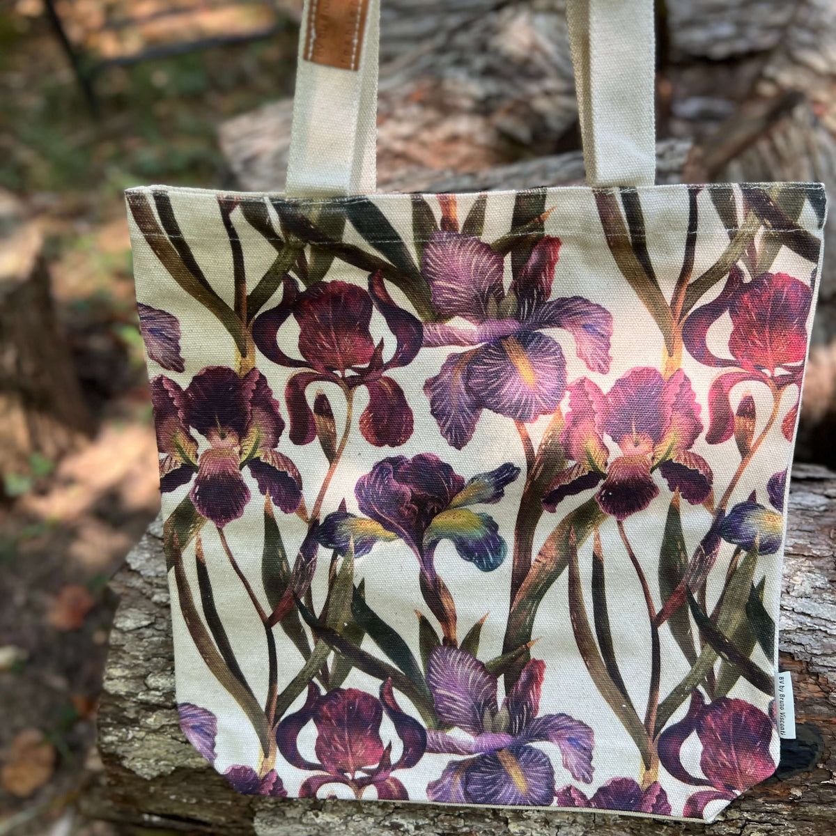 A canvas tote bag with a watercolor-style purple and burgundy iris floral print, featuring natural beige canvas handles, photographed outdoors on a tree trunk with a blurred forest background.
