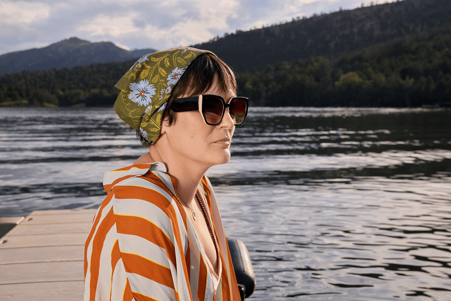 Woman wearing sunglasses and a patterned headscarf by a lake with mountains in the background