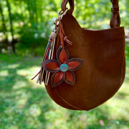 Brown leather handbag with a decorative flower keychain against a blurred green outdoor background