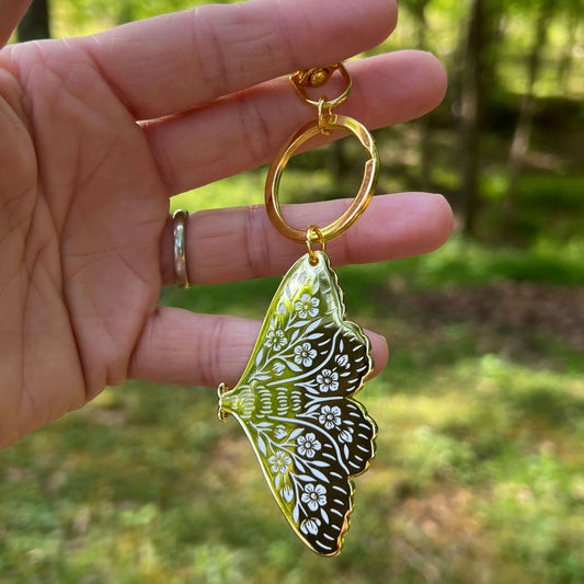 Hand holding a butterfly-shaped keychain with intricate design against a blurred natural background