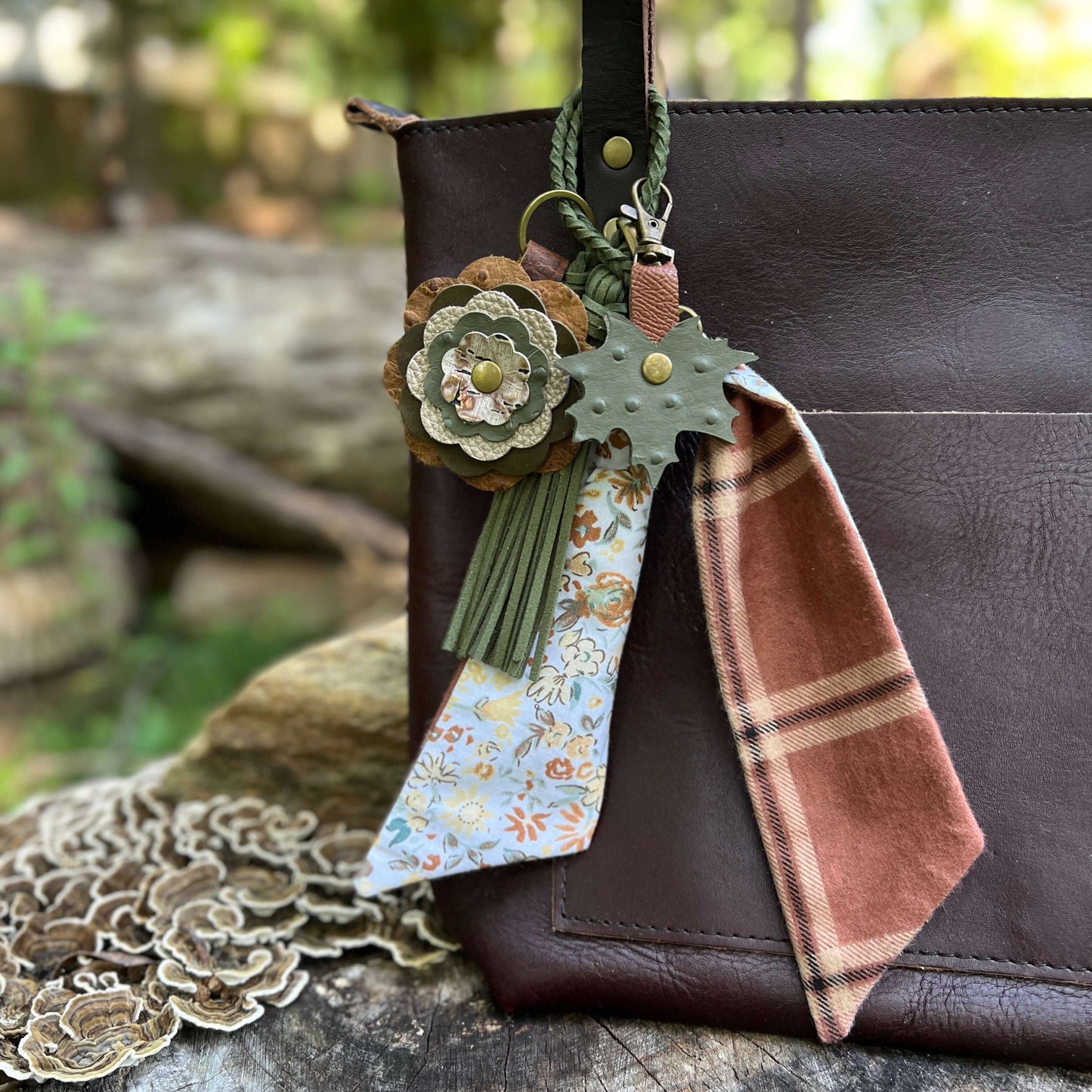 A brown leather shoulder bag with a decorative hanging keychain charm. The charm includes a green leaf-shaped tag, a rust-brown plaid fabric tag, green tassels, and a floral leather flower accent, shown against a blurred outdoor background.