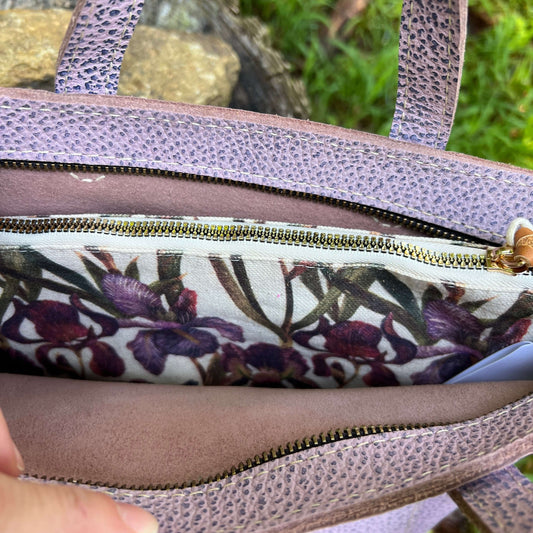 Close-up of a lavender pebbled leather handbag showing the interior lining with a purple and green floral print and a gold zipper.