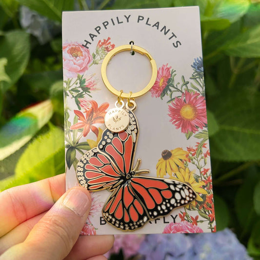 Butterfly keychain on a floral card with 'Happily Plants' branding, held against a natural background.