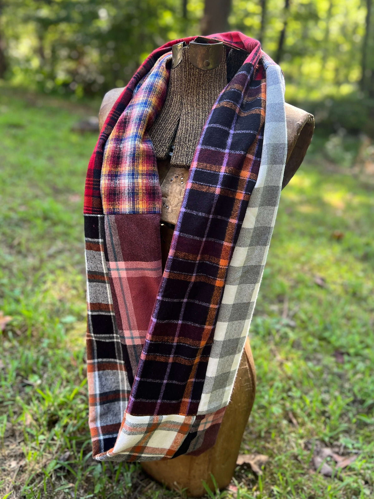 A multicolor patchwork plaid scarf draped on a wooden mannequin torso outdoors on a grassy lawn with trees in the background.