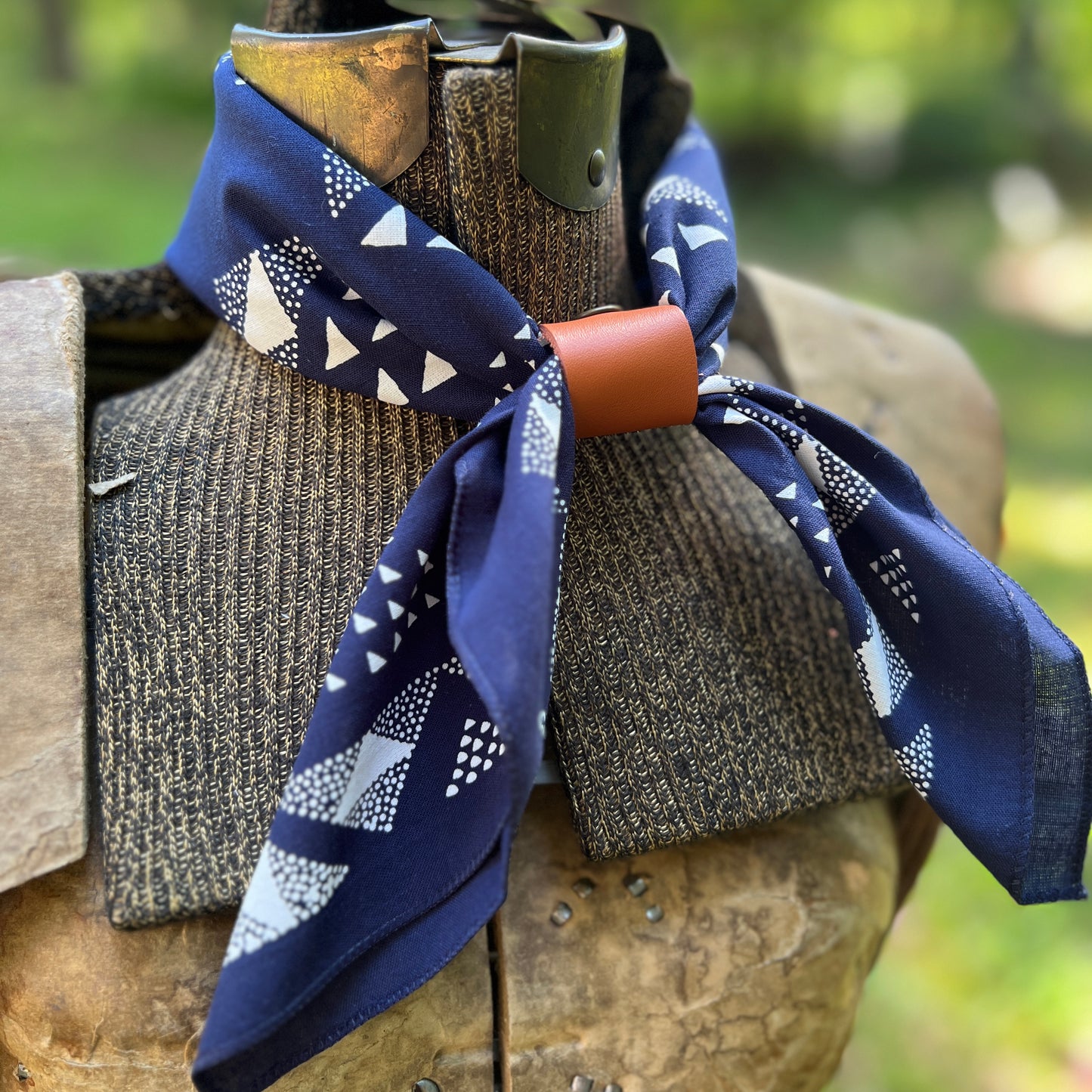 A navy blue patterned neck scarf with white geometric shapes, tied around a rustic brown wooden collar on an outdoor mannequin bust with a blurred green background.
