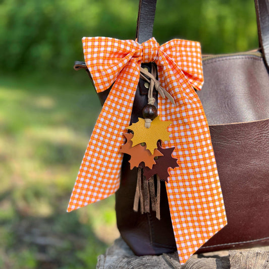 Brown leather bag with an orange checkered bow and leaf charms on a blurred green background