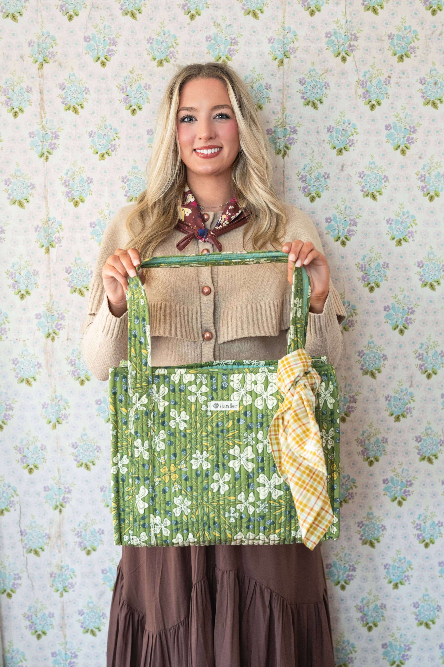 Woman holding a green floral-patterned bag against a floral wallpaper background