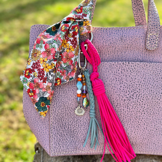 Pink textured handbag with a floral strap and colorful tassel against a blurred green background