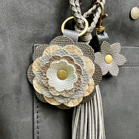 A close-up of a floral leather keychain or bag charm with layered flower petals in beige, taupe, gray, and off-white, gold-tone metal rings and snaps, and a long gray tassel, shown attached to a dark gray leather surface.