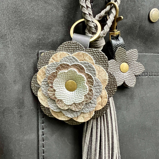 A close-up of a floral leather keychain or bag charm with layered flower petals in beige, taupe, gray, and off-white, gold-tone metal rings and snaps, and a long gray tassel, shown attached to a dark gray leather surface.