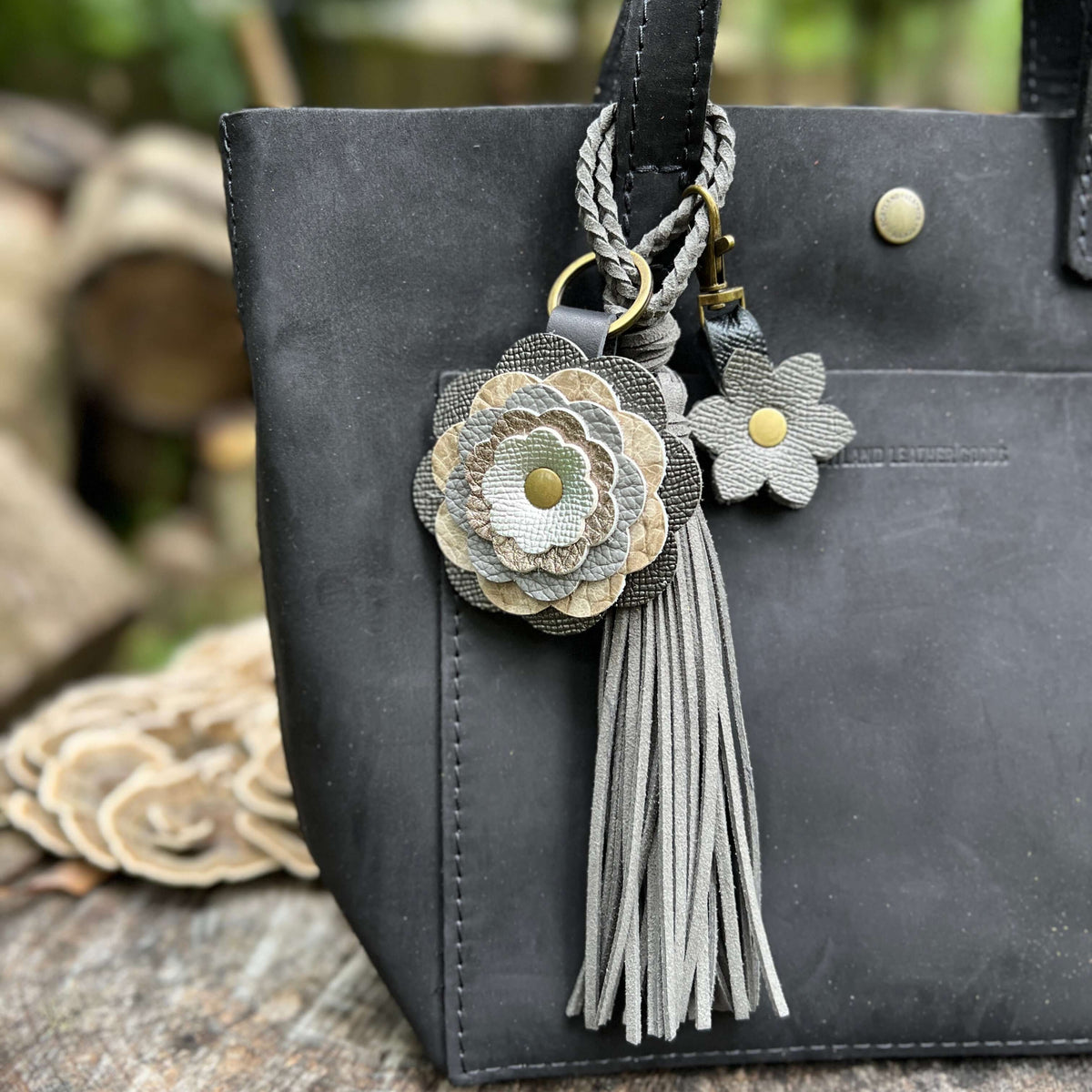 A close-up of a black leather tote bag featuring decorative keychain charms, including a braided strap, a large layered fabric flower in gray and beige tones, a smaller matching flower, and a gray tassel.