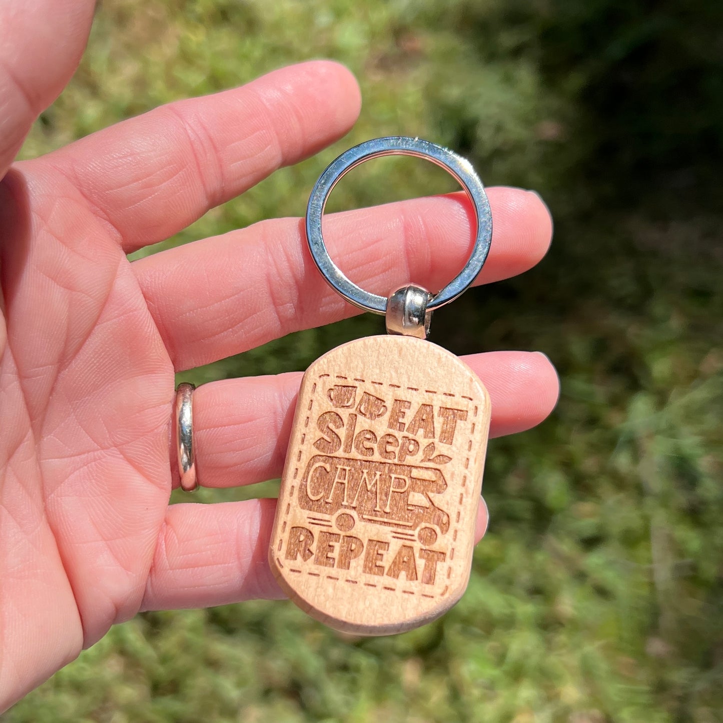 A hand holding a wooden dog-tag style keychain outdoors. The keychain has a silver metal split ring and a light natural wood tag engraved with the phrase 'EAT Sleep CAMP REPEAT'.