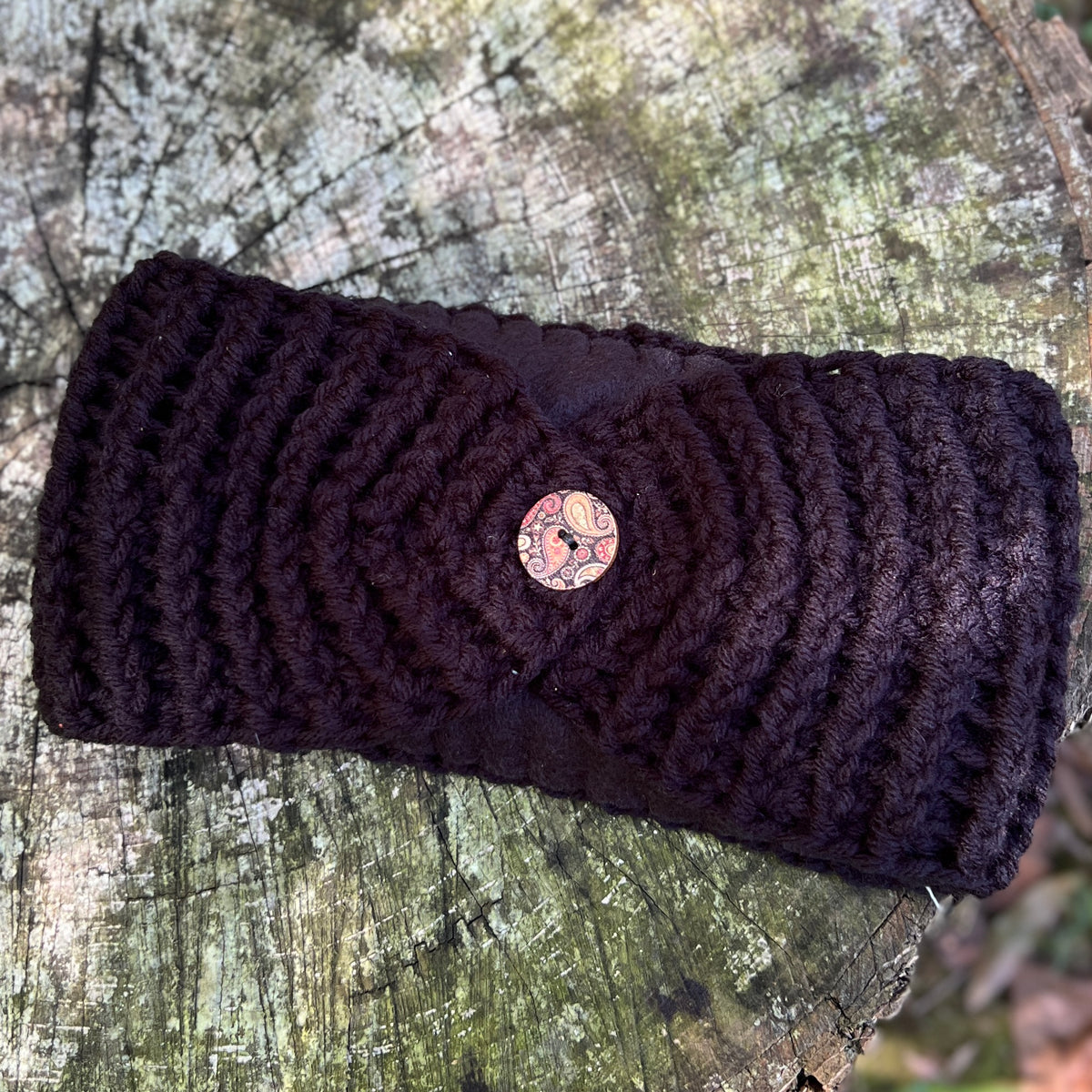 A black crocheted headband with a decorative round button, displayed on a weathered tree stump.