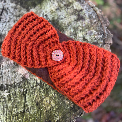 A chunky knit orange-red headband with a brown center panel and a decorative button, displayed on a weathered tree stump.
