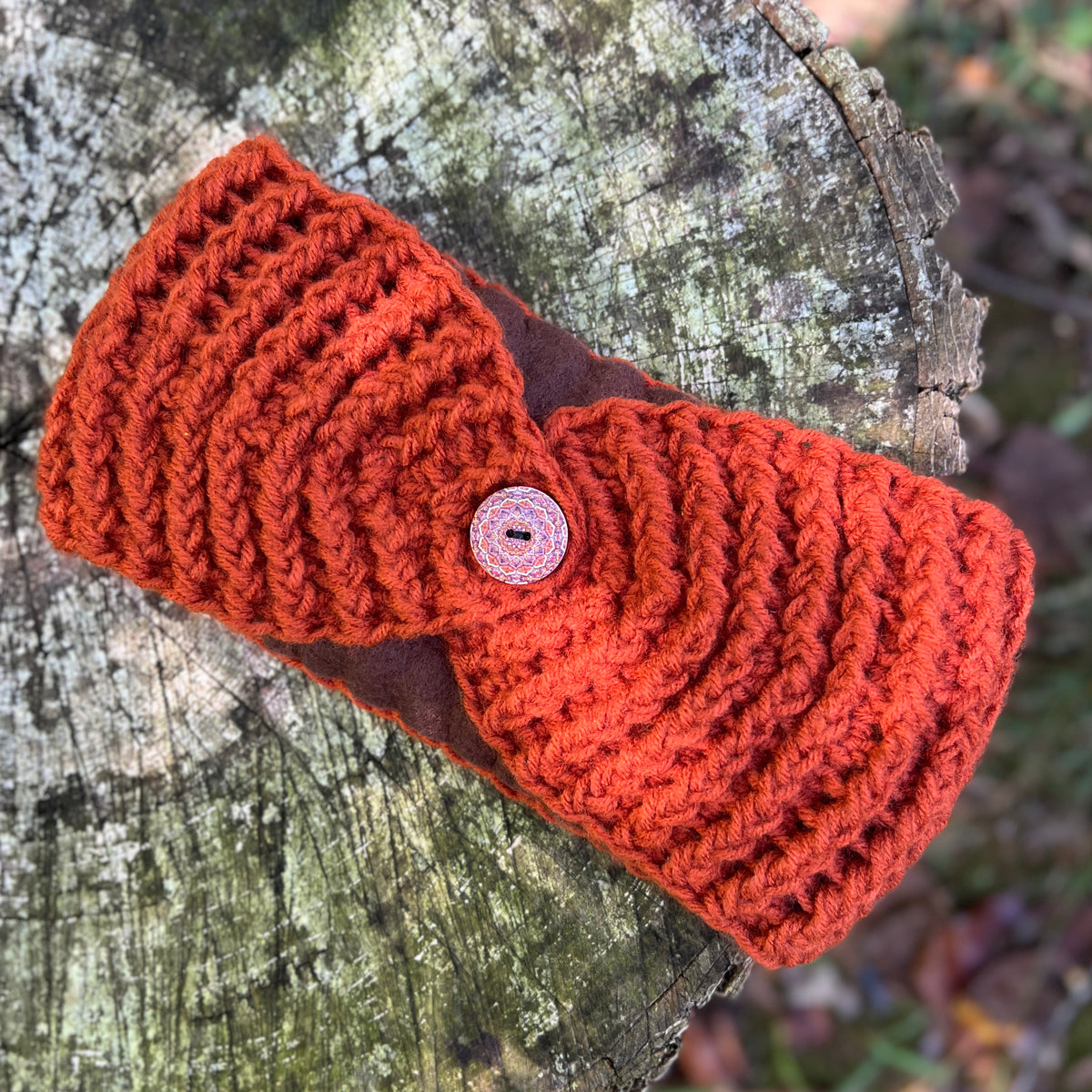 A chunky knit orange-red headband with a brown center stripe and a pink decorative button, displayed on a weathered tree stump outdoors.