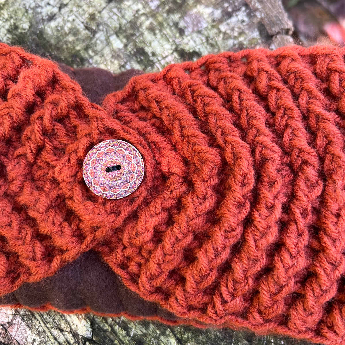 A close-up of a chunky crocheted headband in a rust-orange color, featuring a decorative round button closure and a dark brown inner band, set against a textured outdoor background.