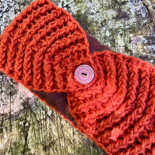 A close-up of a chunky orange-red crocheted headband with a round decorative button, laid flat on a textured, mossy surface.
