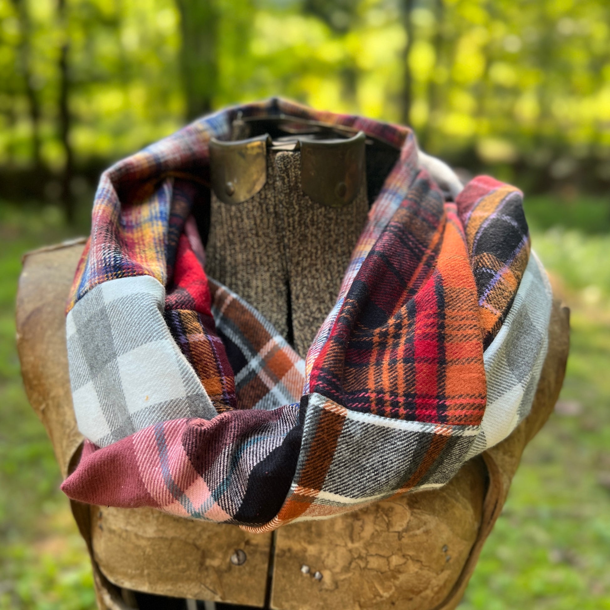 A multicolor plaid infinity scarf draped on a rustic wooden mannequin torso, set against a blurred outdoor forest background.