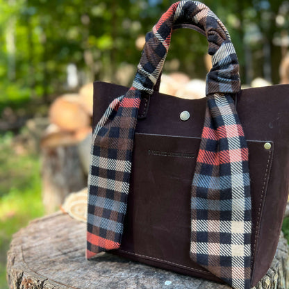 A dark brown leather tote bag with a plaid fabric handle and shoulder strap, resting on a rustic wooden log with a blurred outdoor forest background.