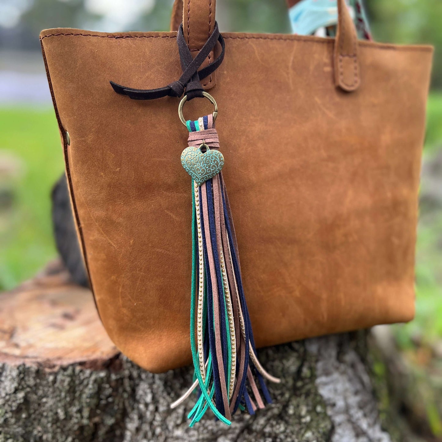 Tassel Purse Charm with Heart CharmChic and simple faux suede tassel purse charm with give your tote or bag an instant boho vibe. Crafted with faux suede in navy, sea green, warm brown and tan with golden studs and accented with heart charm. Long loop wor