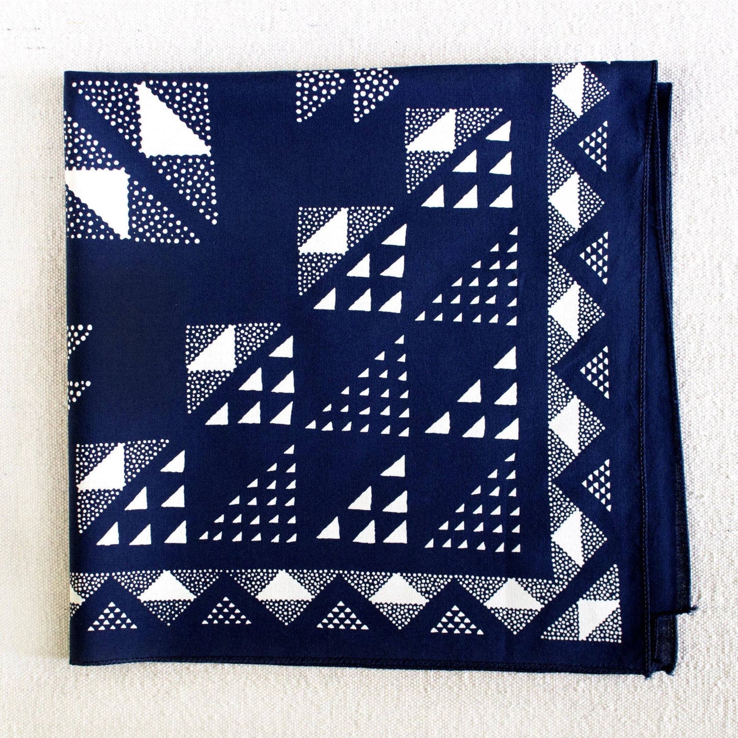 A folded navy blue bandana or pocket square featuring a white geometric pattern of triangles, squares, and dotted lines, laid flat on a textured off-white background.