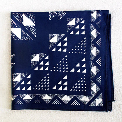 A folded navy blue bandana or pocket square featuring a white geometric pattern of triangles, squares, and dotted lines, laid flat on a textured off-white background.