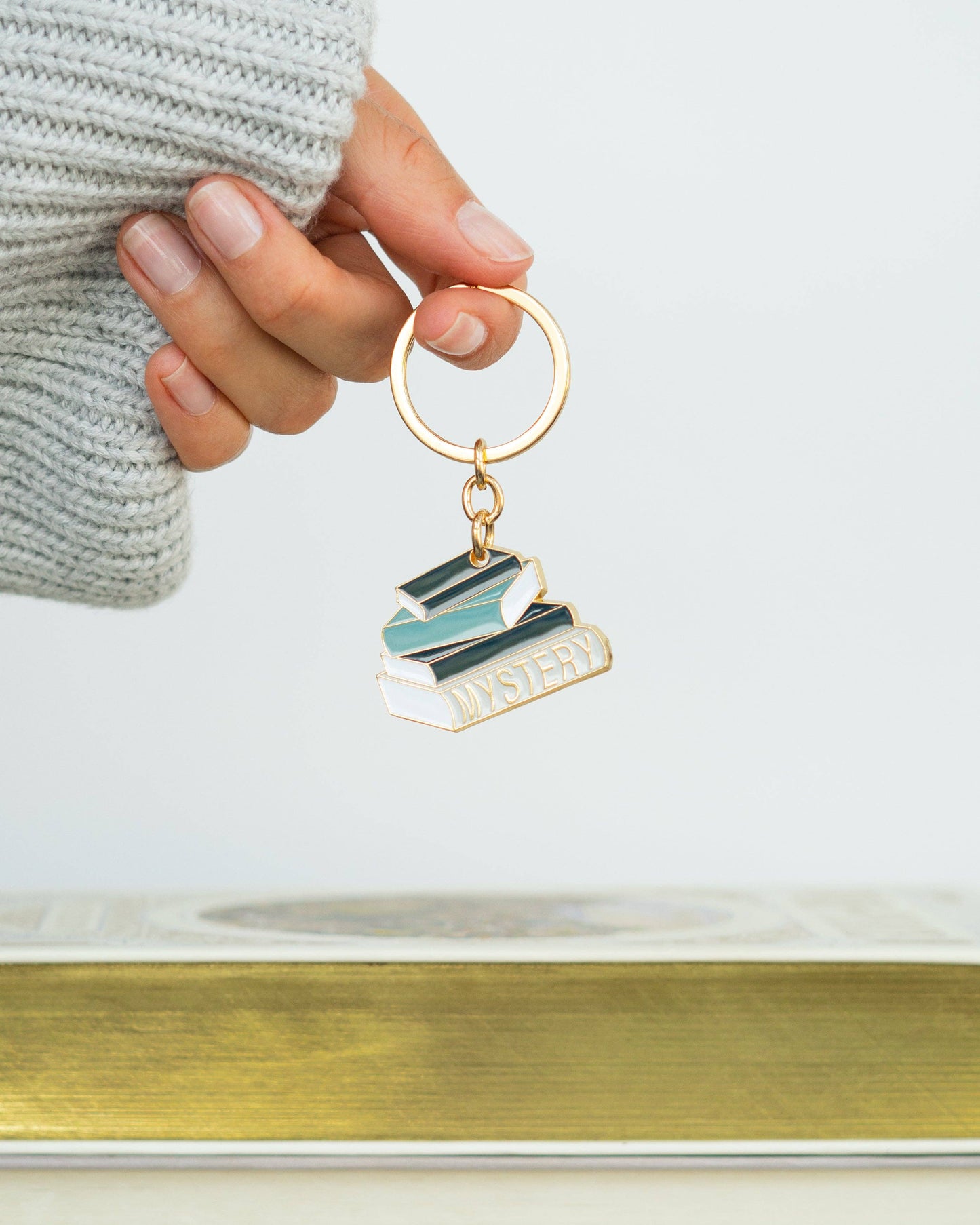 A hand wearing a light gray knit sweater holds a gold-tone keychain charm shaped like stacked books with the word 'MYSTERY' on the front. The background is a clean white studio setting with a blurred gold surface at the bottom.