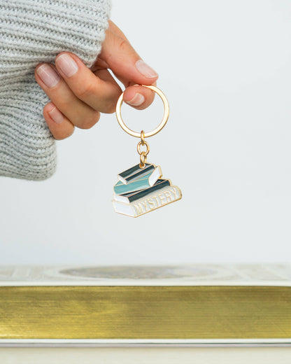A hand wearing a light gray knit sweater holds a gold-tone keychain charm shaped like stacked books with the word 'MYSTERY' on the front. The background is a clean white studio setting with a blurred gold surface at the bottom.