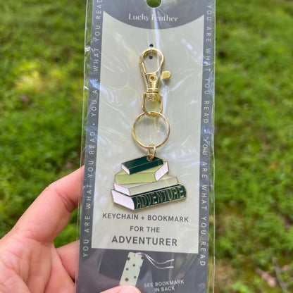 A hand holds a packaged enamel keychain and bookmark set outdoors. The product features a stack of three books with green, white, and dark green enamel details, a gold-tone clasp, and a matching bookmark card in the back.