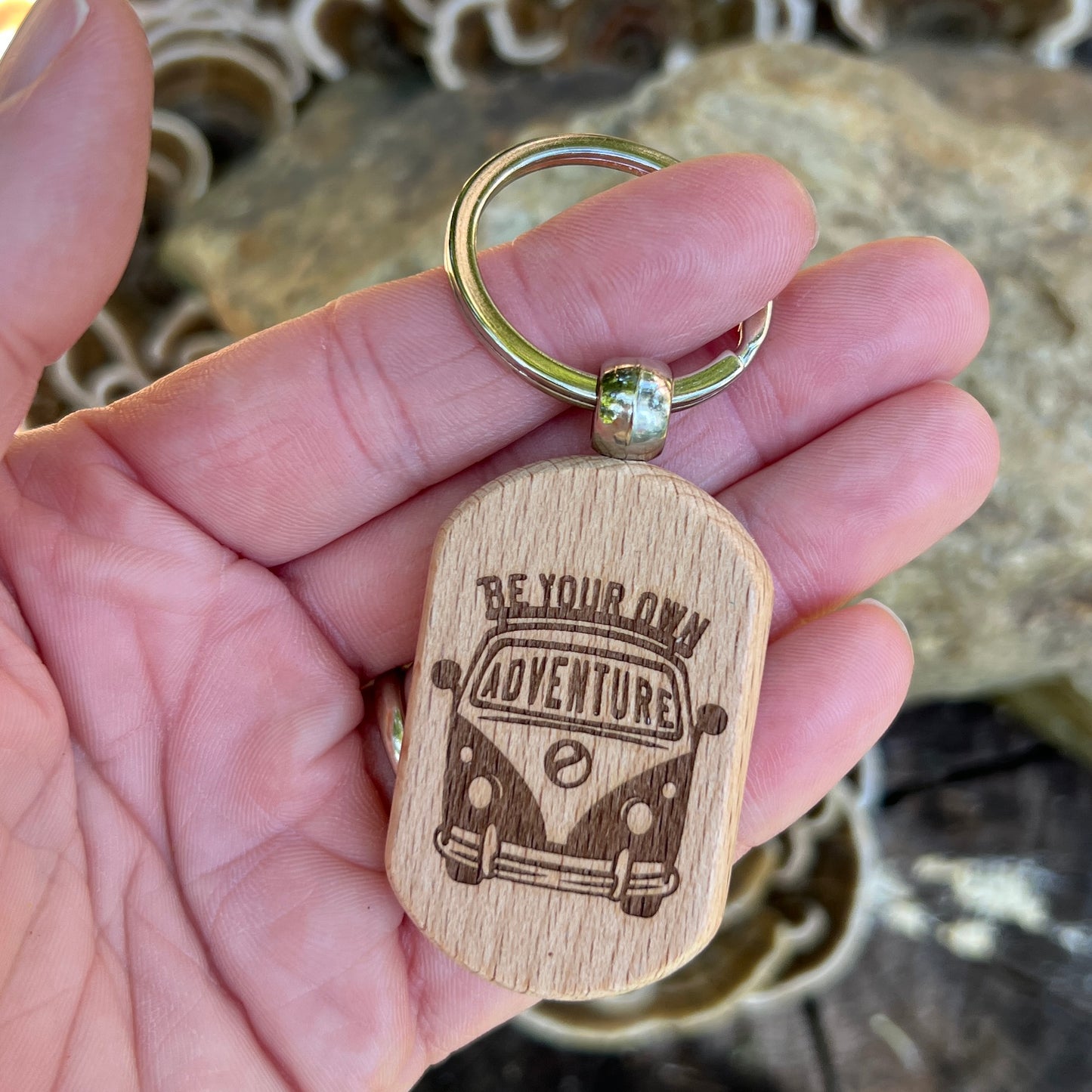 A hand holding a wooden dog tag keychain engraved with a retro VW bus and the words 'BE YOUR OWN ADVENTURE'. The keychain has a silver metal split ring and is photographed against a blurred natural background of rocks and moss.