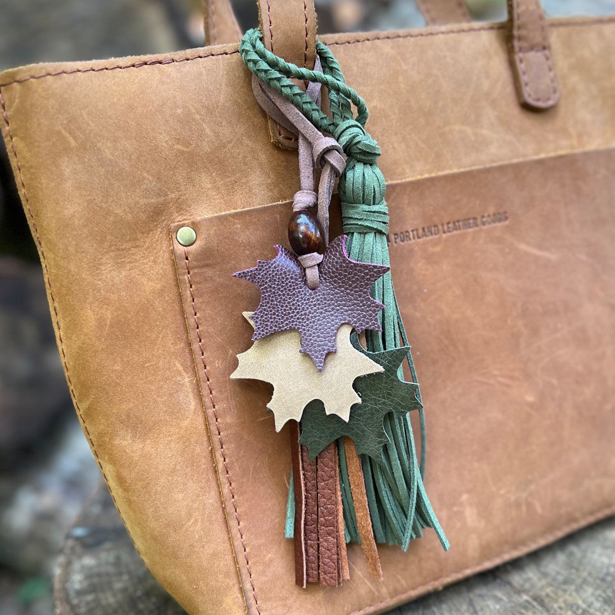 A close-up of a tan leather tote bag with a decorative hanging tassel keychain. The keychain features three layered leaf-shaped leather charms in purple, tan, and dark green, along with braided green cords and tassels.