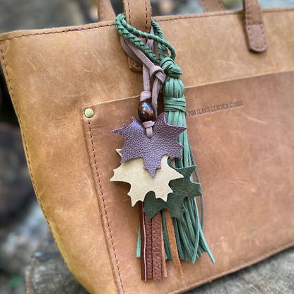 A close-up of a tan leather tote bag with a decorative hanging tassel keychain. The keychain features three layered leaf-shaped leather charms in purple, tan, and dark green, along with braided green cords and tassels.