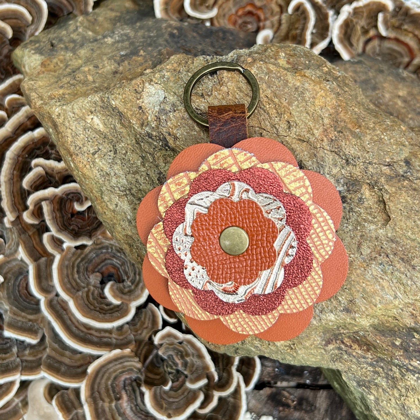 A flower-shaped keychain with layered orange, rust, beige, and white textured leather petals, a brown leather strap, and a brass key ring, displayed on a rock with mushrooms in the background.