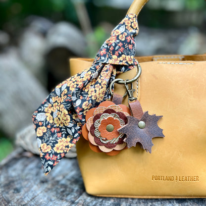 A yellow leather handbag with a floral fabric scarf tied to its handle, featuring a layered autumn flower charm and a leaf-shaped charm. The bag is embossed with the brand name 'PORTLAND A LEATHER' and sits on a rustic wooden surface with a blurred outdoor background.