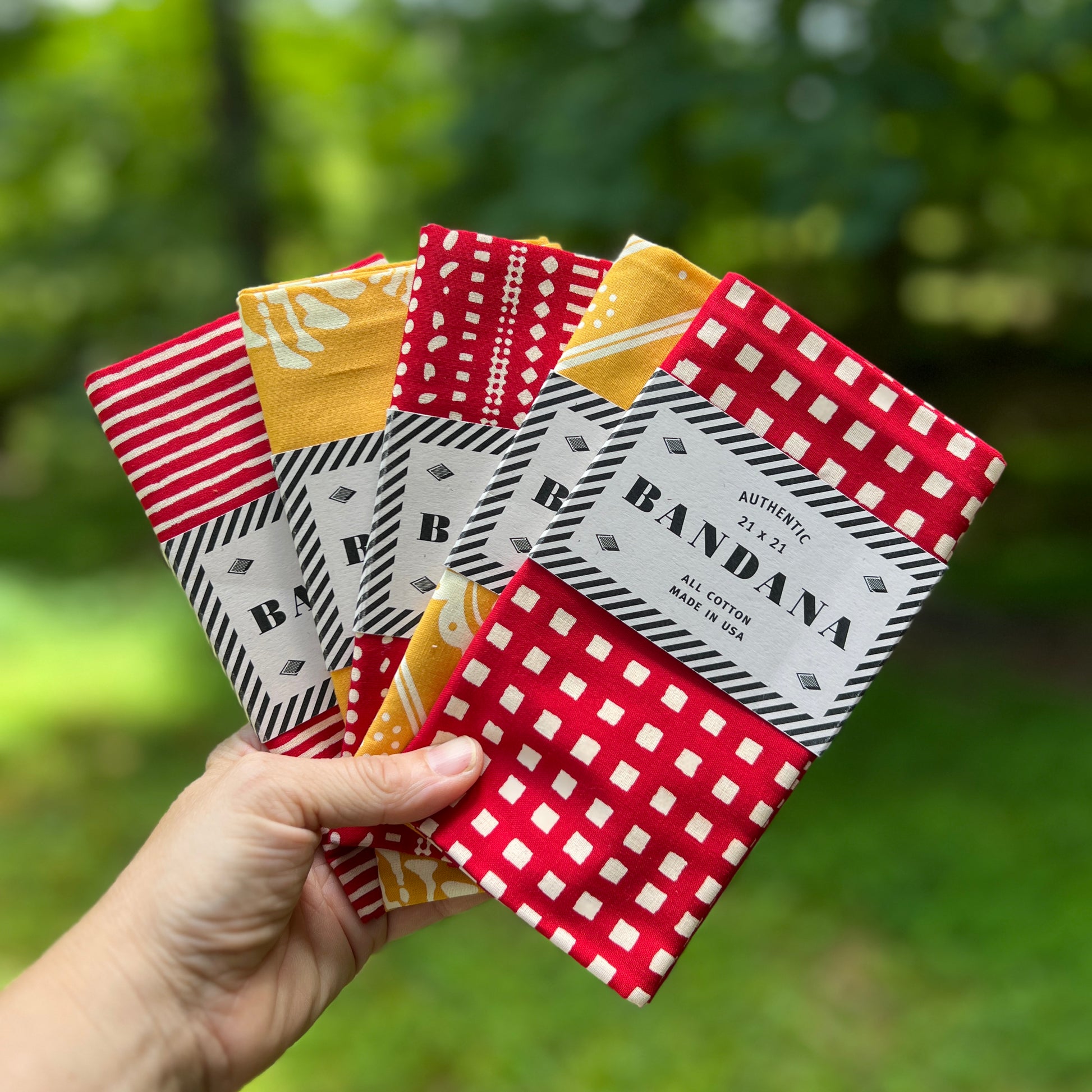 Hand holding a collection of patterned bandanas with 'B' brand label against a blurred green background