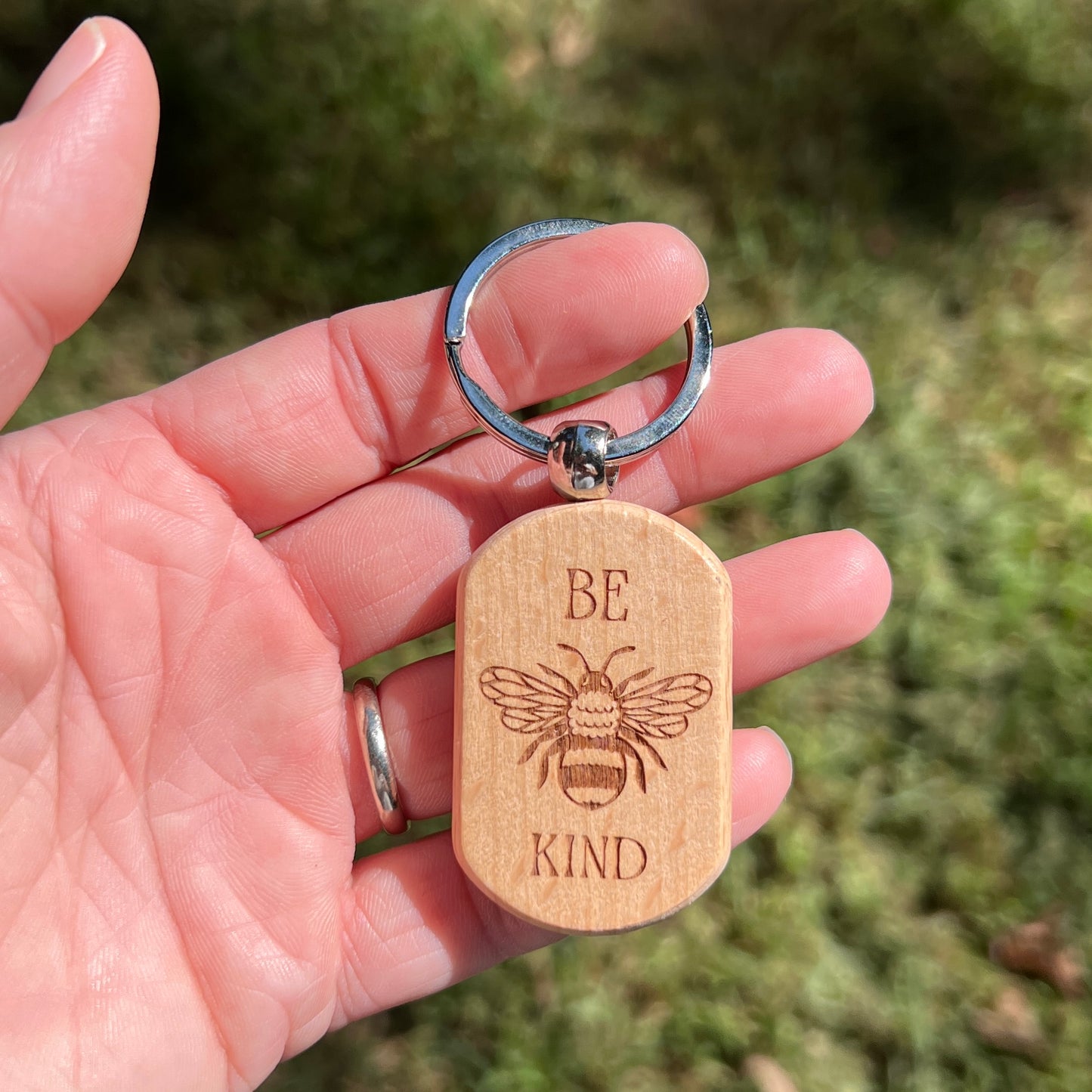 A hand holding a wooden keychain with a silver split ring. The wooden tag is engraved with a bee and the words 'BE KIND'.