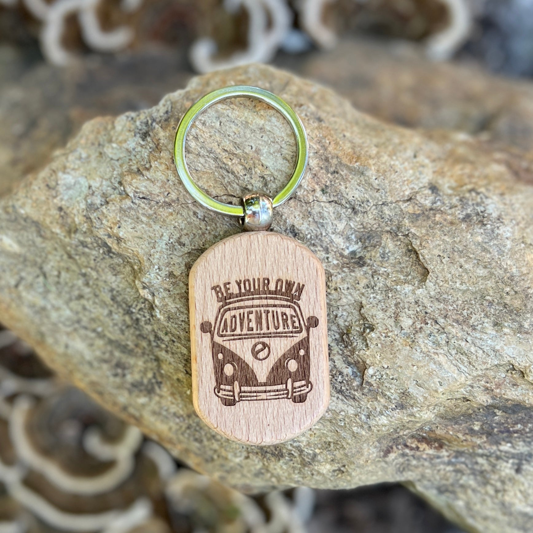 A wooden dog-tag style keychain with an engraved van design and the words 'BE YOUR OWN ADVENTURE' hanging from a silver key ring, displayed on a textured rock.