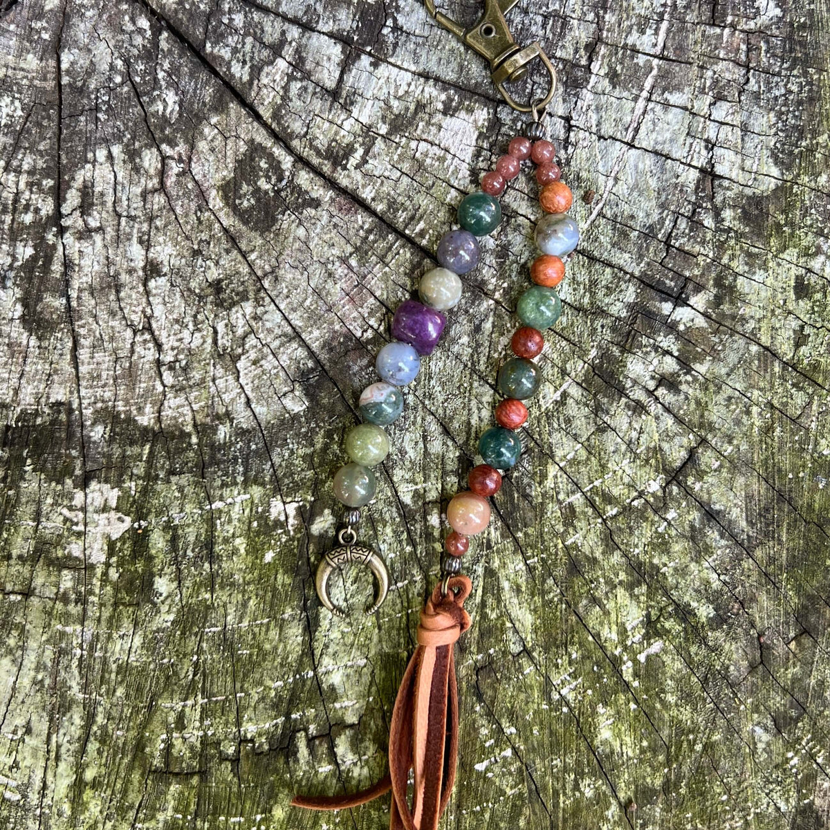 A beaded keychain  with multicolored stone beads, a bronze crescent moon charm, and a brown leather tassel, displayed on a weathered wood surface.