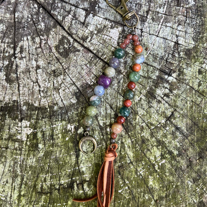 A beaded keychain  with multicolored stone beads, a bronze crescent moon charm, and a brown leather tassel, displayed on a weathered wood surface.