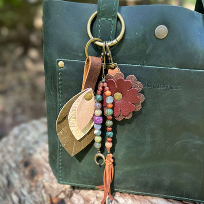 A leather keychain bag charm set featuring a dark green leather bag, a brown leather flower, two leaf-shaped leather pieces, colorful beaded strands, and a tassel, shown outdoors on a tree trunk.