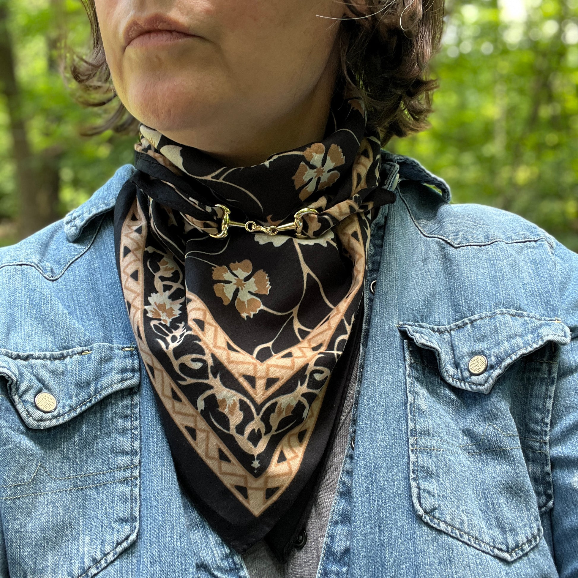 A close-up of a person outdoors wearing a black and beige floral bandana scarf tied around their neck, paired with a blue denim jacket.