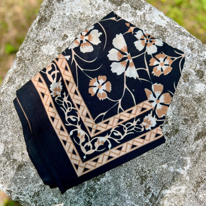 A folded black bandana with a white and tan floral print and geometric border, displayed on a textured gray stone outdoors.